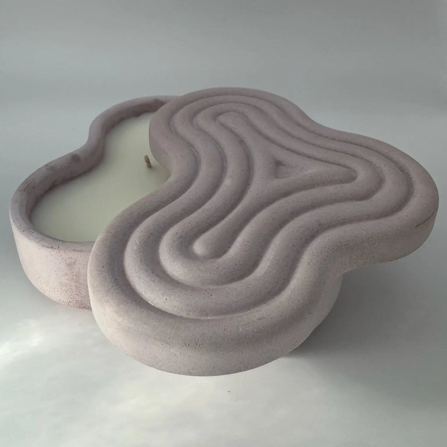 A light purple concrete candle jar with two wicks, its lid partially covering the jar to reveal the wicks. The candle is heart-shaped and rests on a white background with a subtle overcast shadow, adding to its aesthetic appeal.