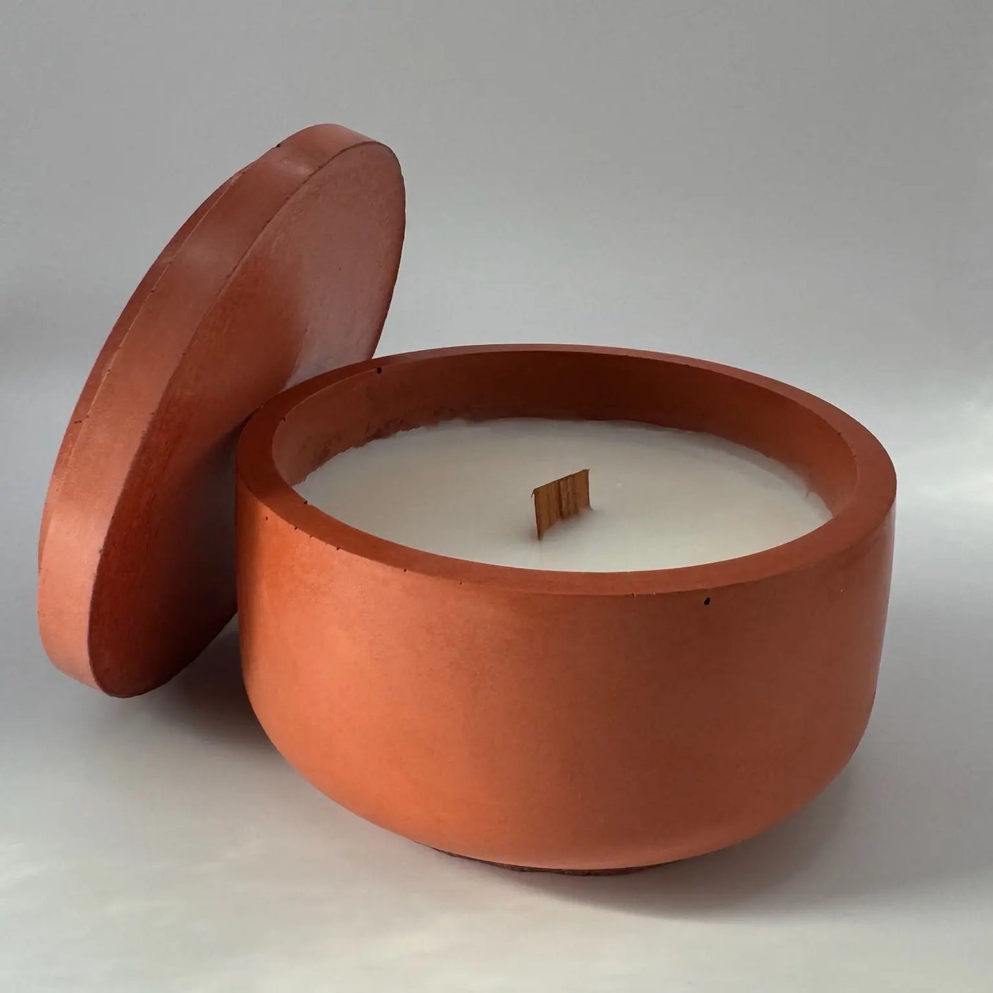 A rust terracotta concrete candle jar with a wood wick, accompanied by its lid leaning against the jar in the foreground. The candle rests on a white background with a subtle shadow, enhancing its aesthetic appeal.