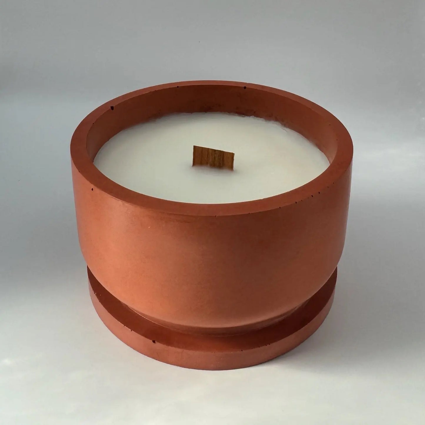 A rust terracotta concrete candle jar with a wood wick, resting on its jar lid. The candle rests on a white background with a subtle shadow, enhancing its aesthetic appeal.