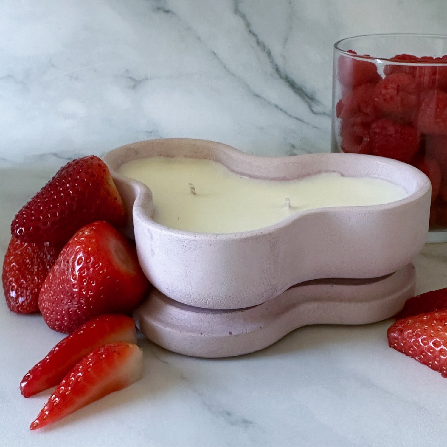 A heart-shaped light purple concrete candle jar with two cotton wicks, resting on its lid. Surrounding the candle are fresh strawberries and a jar of raspberries against a white-grey marble background.
